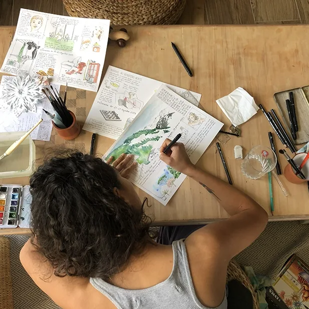 Vue de dessus : Débora Borba dessine dans un carnet aquarellé, entourée de pinceaux, d'une boîte d'aquarelle et de planches déjà peintes