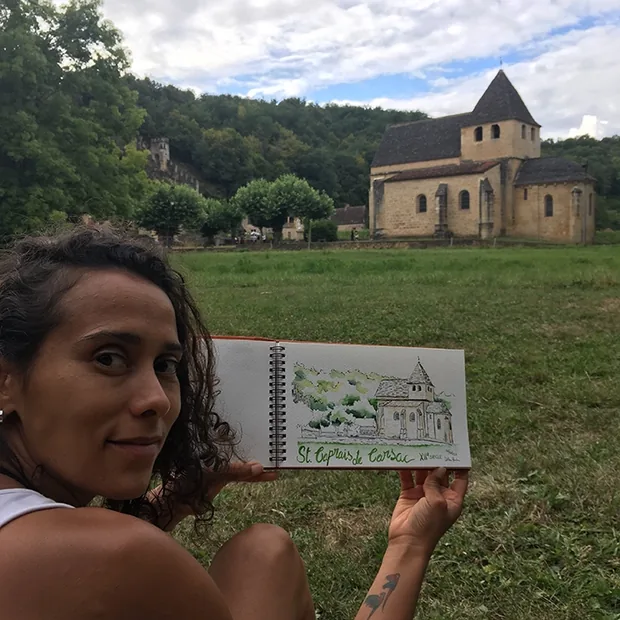 Débora Borba tient son carnet de croquis devant une église romane, le dessin reprenant le paysage derrière elle
